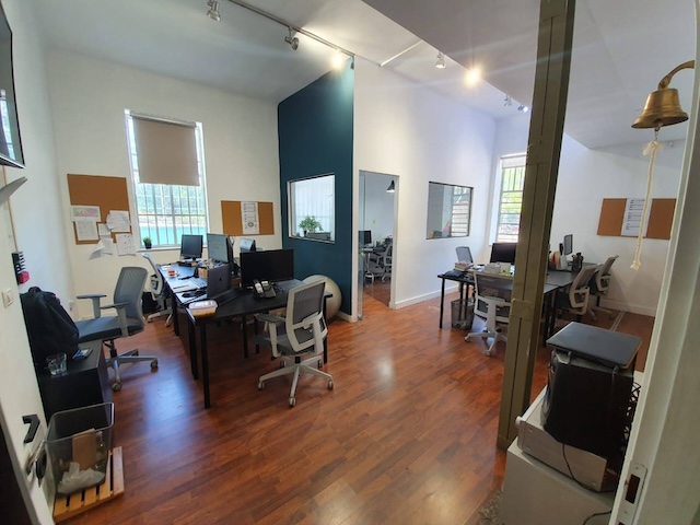 משרד להשכרה מודרני בעל חלל פתוח עם מספר שולחנות, כיסאות משרדיים, מחשבים, לוחות שעם על הקירות, רצפת עץ ופעמון התלוי ליד הכניסה. המשרד ממוקם ליד איסרליש ומונטיפיורי בתל אביב, והוא מואר היטב באור טבעי.